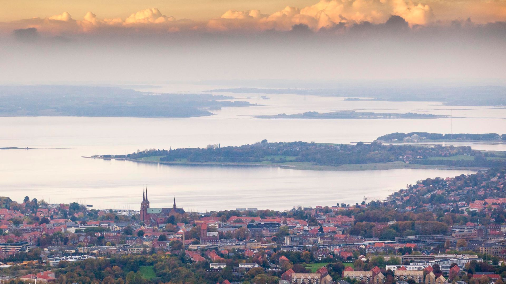 Udsigt over Roskilde med Roskilde Domkirke i forgrunden og Roskilde Fjord omgivet af øer og landskab i baggrunden under en himmel med skyer i aftensol.