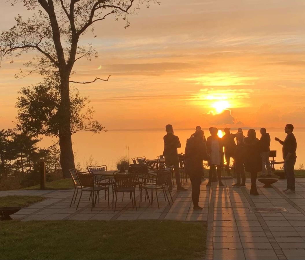 Gruppe mennesker står på terrassen ved Bautahøj Hotel & Konferencecenter og nyder en smuk solnedgang over vandet.