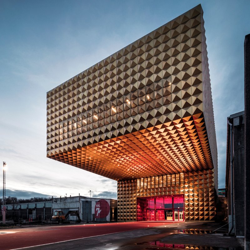 Gylden, kubeformet facade på Rockmuseet med markant udhæng og rødlyst indgang i aftenskumring.