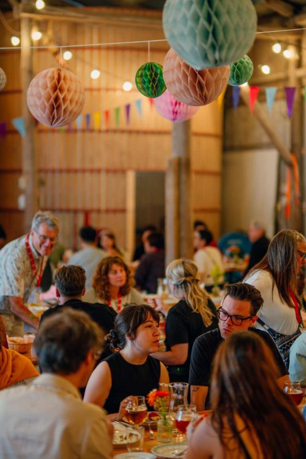 Gæster sidder ved langborde i en rustik lade hos Herslev Bryghus, omgivet af farverige papirdekorationer og hyggelig belysning.