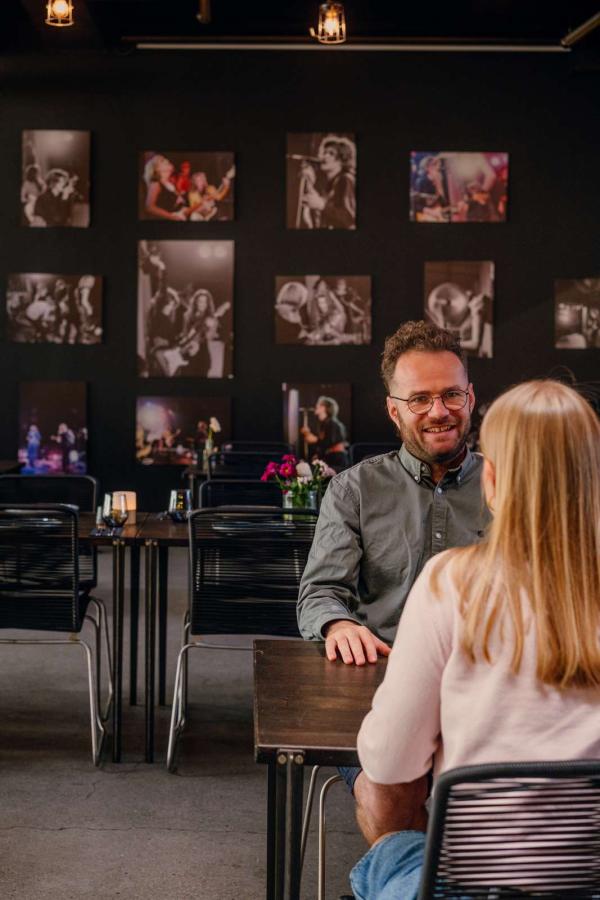 To personer sidder ved et bord i Rockmuseets café, omgivet af vægge dekoreret med rockkoncertbilleder.